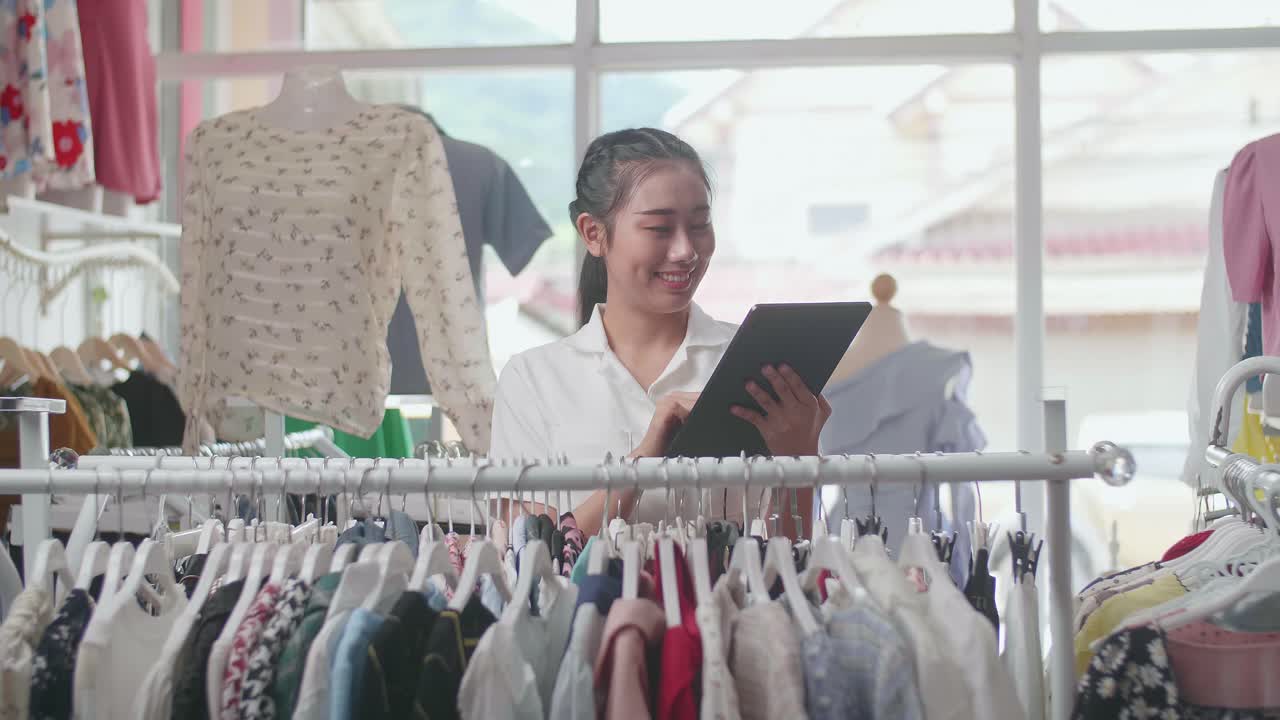 라인 에 있는 재고 를 확인 하기 위해 디지털 태블릿 을 사용 하는 동안 카메라 에 미소 짓는 의류 가게 의 아시아인 여성 소유자
