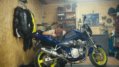 a short-haired man in a gray t-shirt repairs and inspects his blue motorcycle in the garage, a workshop that is equipped with tools and protective clothing