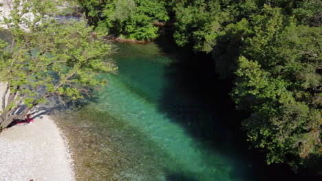 A-woman-enjoys-the-sun-while-lying-on-the-pebbly-riverbank-of-the-famous-Voidomatis-River-that-flows-through-the-Vikos-National-Park-in-Epirus,-Greece