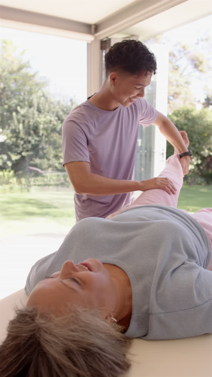 Vertical video: Stretching senior woman's leg, physiotherapist working during therapy session indoor