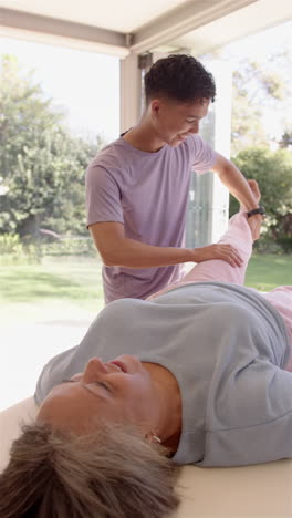 vertical video: stretching senior woman's leg, physiotherapist working during therapy session indoor