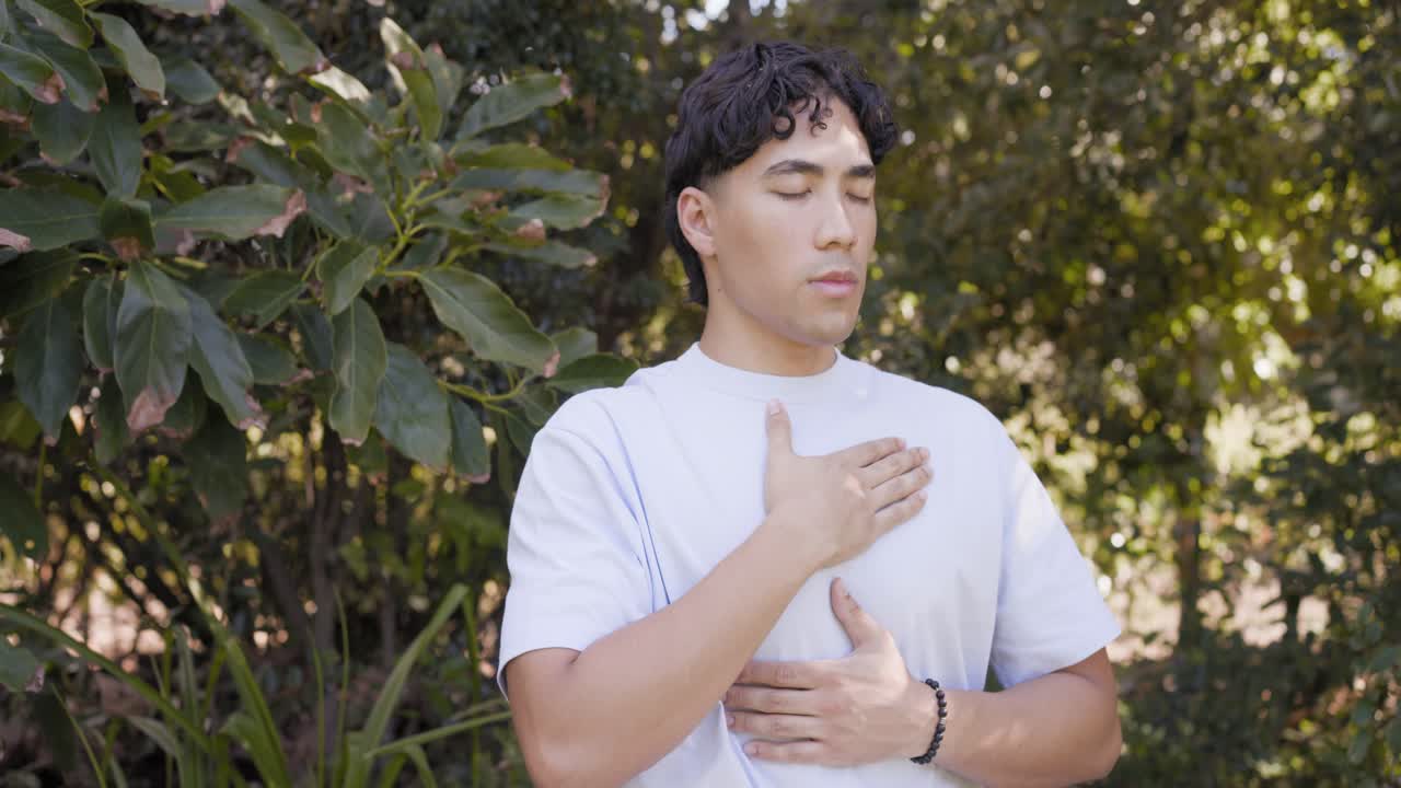 Uomo asiatico in piedi con gli occhi chiusi, che respira, con le mani sul petto e sull'addome, praticando una respirazione calma in giardino.