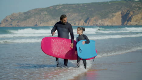 long shot of surfer with artificial leg and daughter holding surfboard and talking while walking on the beach