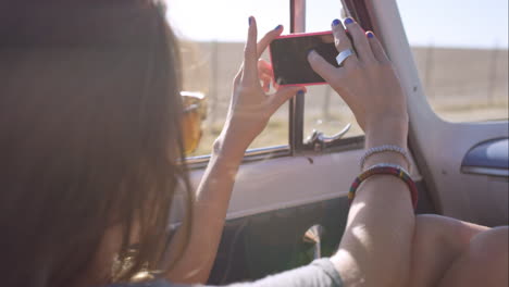 beautiful young woman taking photos with smart phone on road trip in convertible vintage car