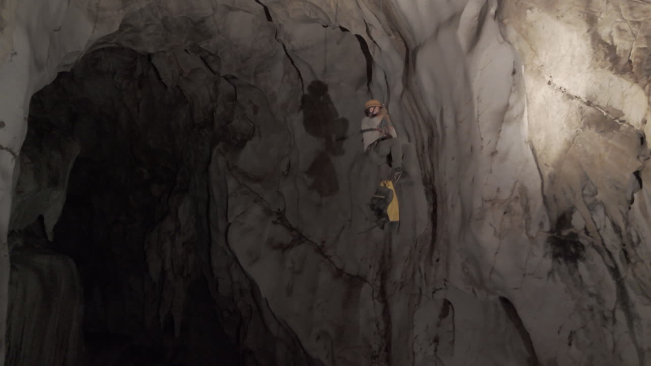 A caver uses Single Rope Technique to control a yellow haul bag along the massive rock wall of Anxiety State Cave in Chiang Mai, Thailand. Adventure, climbing, or exploration themes.