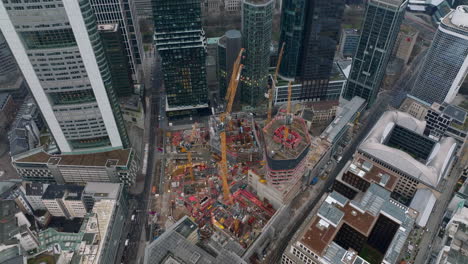 High-angle-shot-of-construction-site-of-new-building.-Orange-tower-cranes-and-machinery.-Tilt-up-reveal-of-downtown-skyscrapers.-Frankfurt-am-Main,-Germany