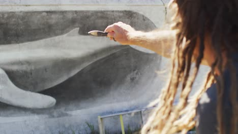 video of caucasian male artist with dreadlocks measuring proportions of whale mural with paintbrush