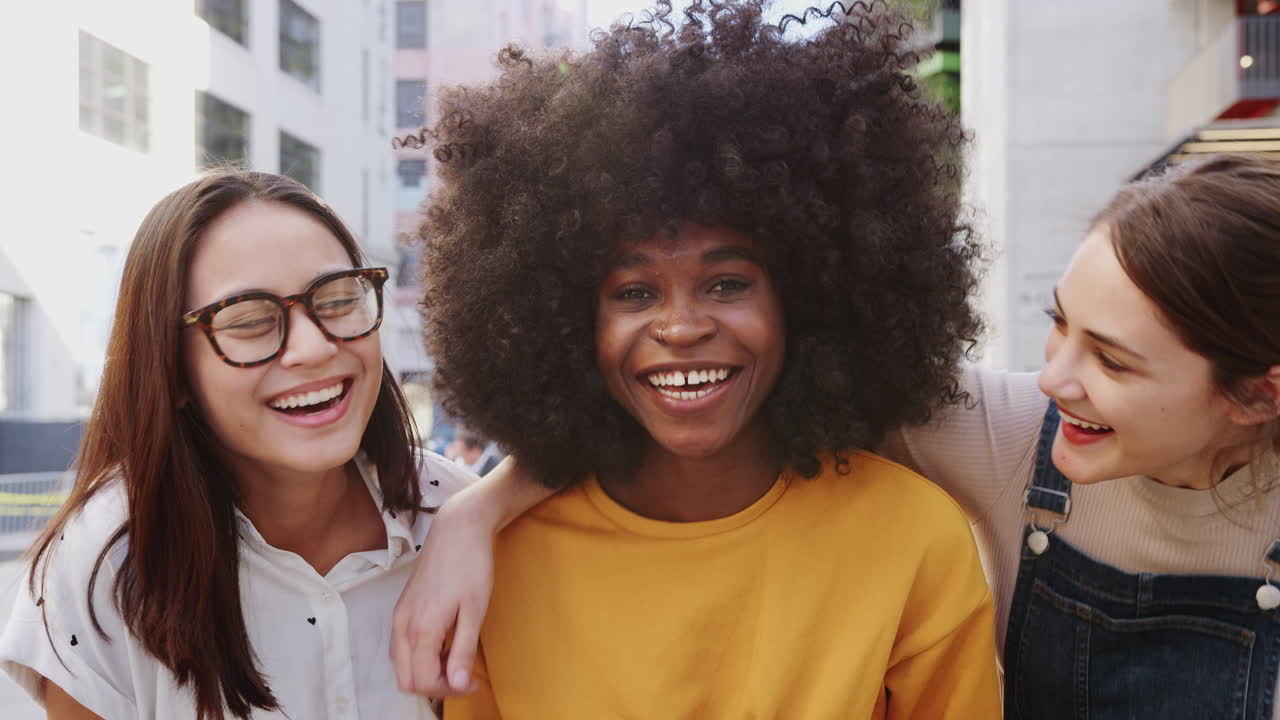 Three young adult girlfriends standing together in a city street laughing to camera, close up