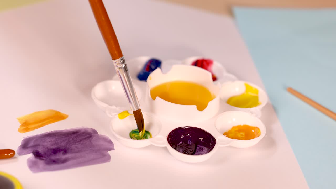 Hand mixes green paint on white palette using brush, close-up, warm indoor lighting, creative workspace