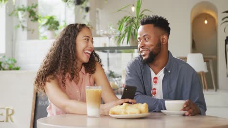 Happy-diverse-couple-spending-time-together-at-cafe,-drinking-coffee-and-using-smartphone