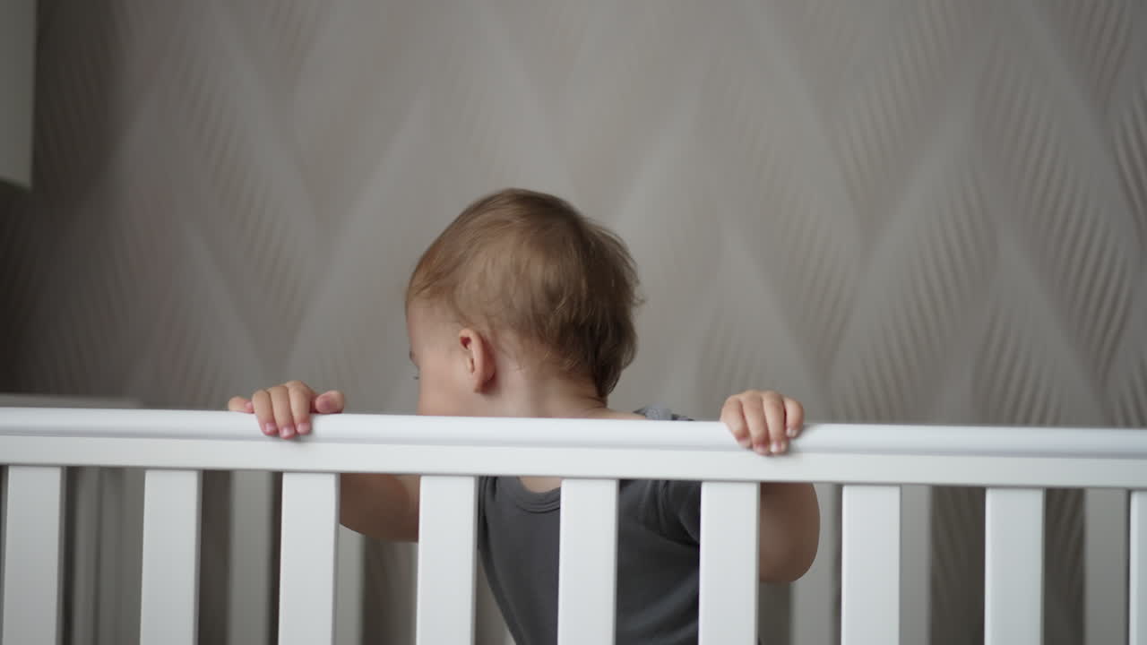 Little boy standing up and sitting down in the cot. Adorable Caucasian child being active after waking up.