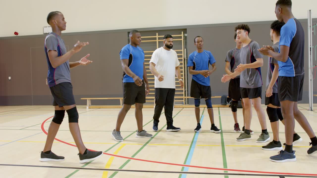 Trainer spornt diverses männliches Volleyballteam an, das sich am Netz in der Sporthalle versammelt und vor dem Spiel die Hände hebt.