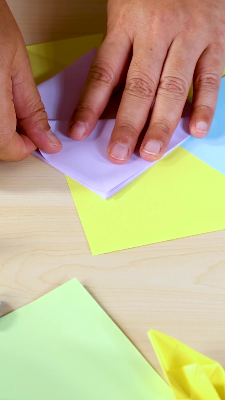 Una persona piega carta origami pastello su un tavolo di legno chiaro sotto un'illuminazione brillante e uniforme, in primo piano.