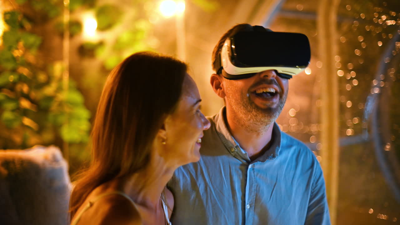 A hugging couple inside transparent bubble tent at glamping at night while man using VR glasses. Illumination inside