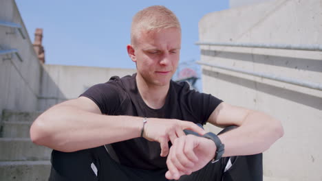 young blond man in workout clothes uses phone and smartwatch outdoors