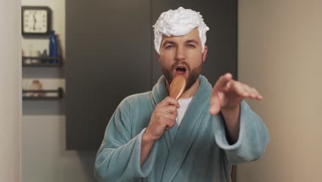 a man with a foam hairdo sings into the brush wearing a bathrobe