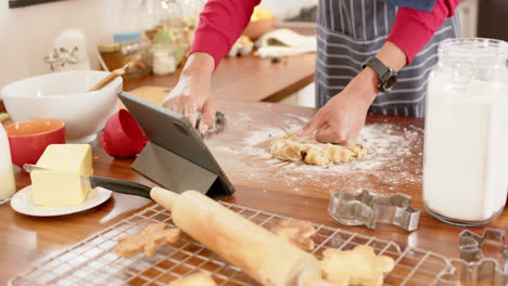 Biracial-man-wearing-christmas-hat,-making-christmas-cookies-using-tablet-at-home,-slow-motion