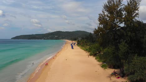 pedestal drone shot of the length of karon beachfront showing the scenic beauty of a sandy beach with crystal-clear waters, against a backdrop of blue skies and lush green trees, in phuket, thailand