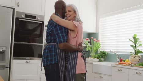 happy senior diverse couple wearing aprons and embracing in kitchen