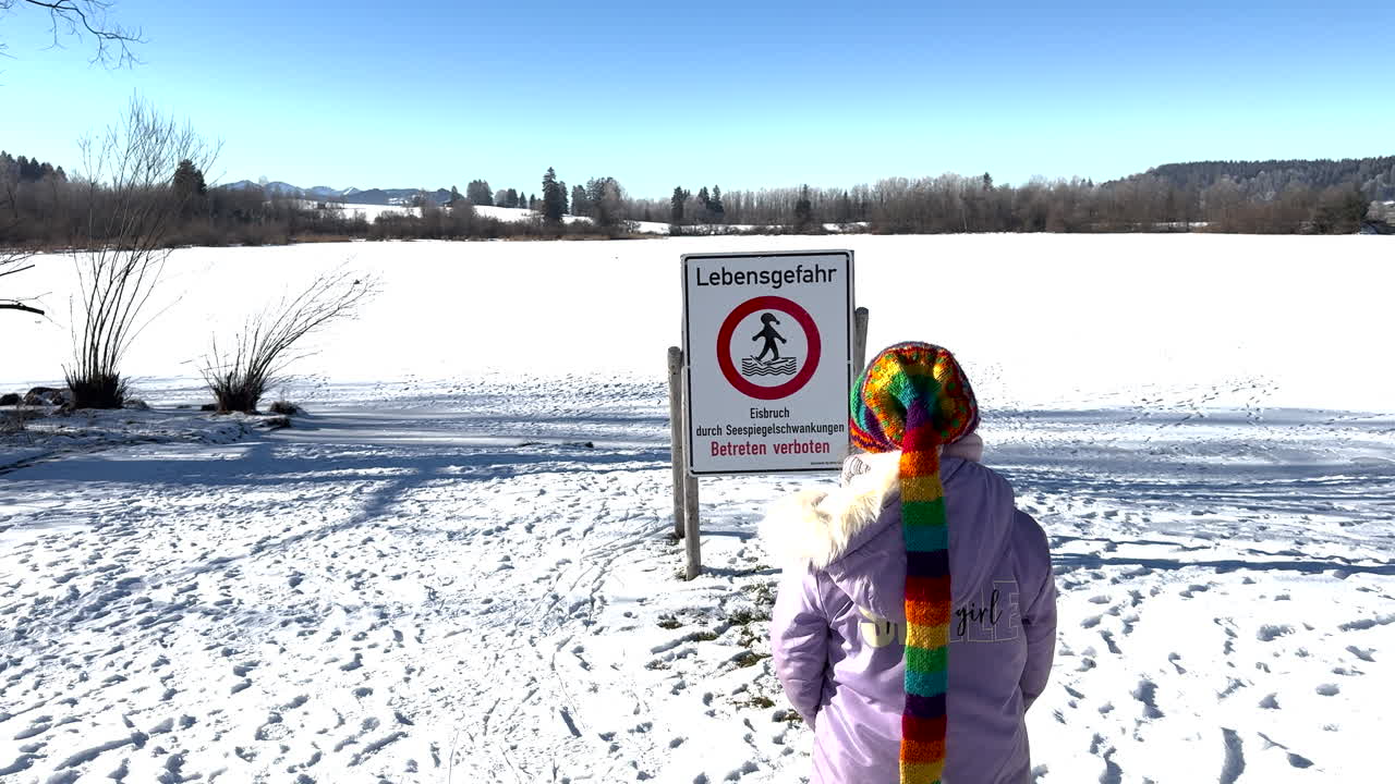 A humorous moment as a child in a colorful scarf unintentionally walks into a "Danger to Life" sign near the frozen Rottachsee. Snowy landscape with bare trees, bright blue sky. Rottachsee, Germany
