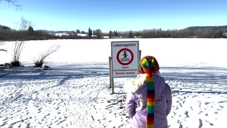 A humorous moment as a child in a colorful scarf unintentionally walks into a "Danger to Life" sign near the frozen Rottachsee. Snowy landscape with bare trees, bright blue sky. Rottachsee, Germany