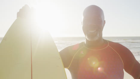 Portrait-of-happy-senior-african-american-man-holding-surfboard-at-beach,-in-slow-motion