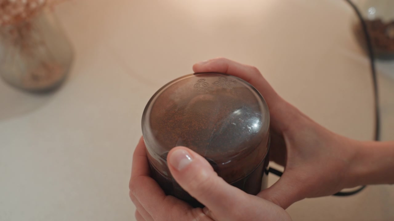 Primer plano de manos sosteniendo un molinillo de café con iluminación suave y cálida. Barista casero preparando granos, rotaciones suaves, uñas pulidas, jarrón de cerámica al fondo, encimera de cocina acogedora, movimiento táctil.