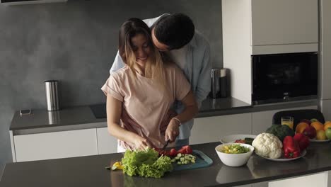 a young man is kissing his girlfriend when she's making a fresh salad for them. he came to hug her and appreciate. long haired woman kisses him back. modern kitchen in grey colours