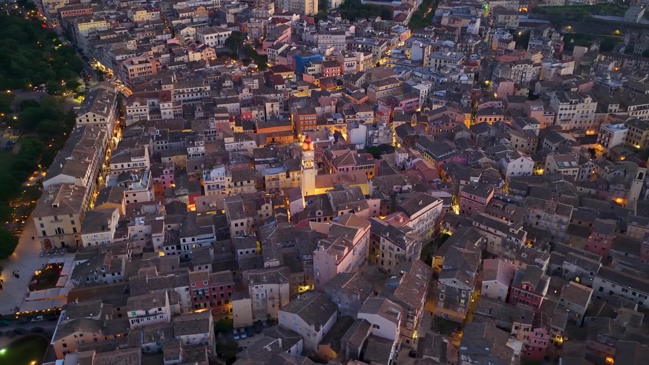 Vista aerea con drone della storica città vecchia di Corfù al crepuscolo con strade illuminate, architettura tradizionale e l'iconico campanile della chiesa di San Spiridione che risplende la sera.