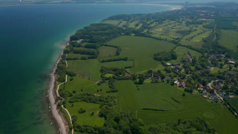 Amazing-high-aerial-view-of-green-countryside-fields-facing-turquoise-Baltic-sea,-tilt-up,-sunny-day