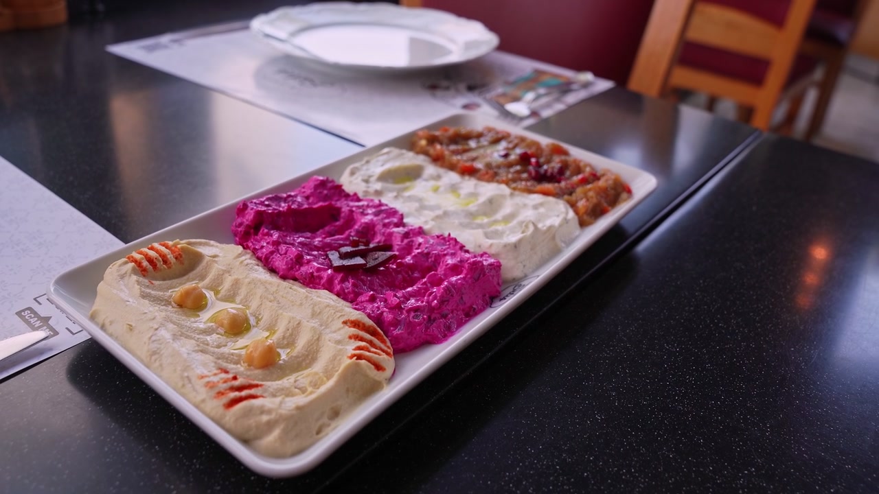 Una ripresa ravvicinata di un piatto bianco con quattro tipi di salse mediterranee, tra cui hummus, crema di barbabietola e moutabal, servito su un tavolo nero in un ambiente da ristorante
