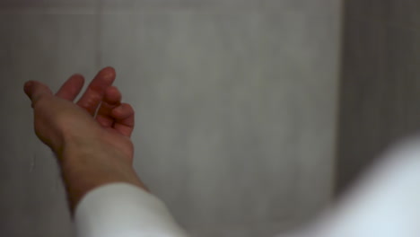 Over-the-shoulder-shot-of-the-hand-of-a-man-wearing-long-sleeves-white-shirt-feeling-the-water-temperature-in-the-shower