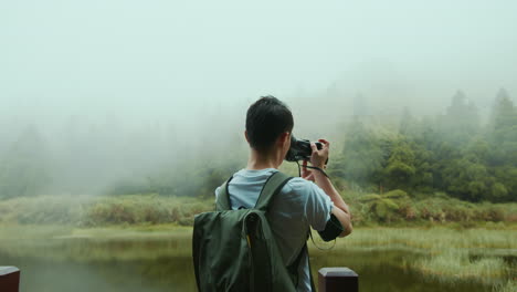 a young asian male photographer in a white t-shirt and a backpack is taking photographs of nature by a lake in the mountains, in taiwan