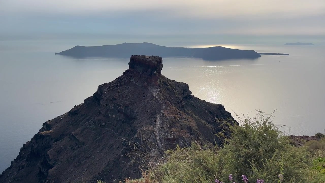 Sweeping wide landscape shot of Skaros Rock in Imerovigli, Santorini, overlooking the calm Aegean Sea at golden hour, with dramatic coastline, serene waters, and tranquil natural lighting.