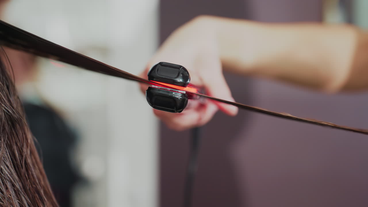 미용실에서 젖은 머리를 한 백인 고객에게 스타일리스트가 단계별 튜토리얼로 머리를 펴는 모습을 클로즈업한 장면입니다. 손, 머리카락의 장력, 매끄럽게 펴는 리듬, 교육적인 분위기가 강조됩니다.