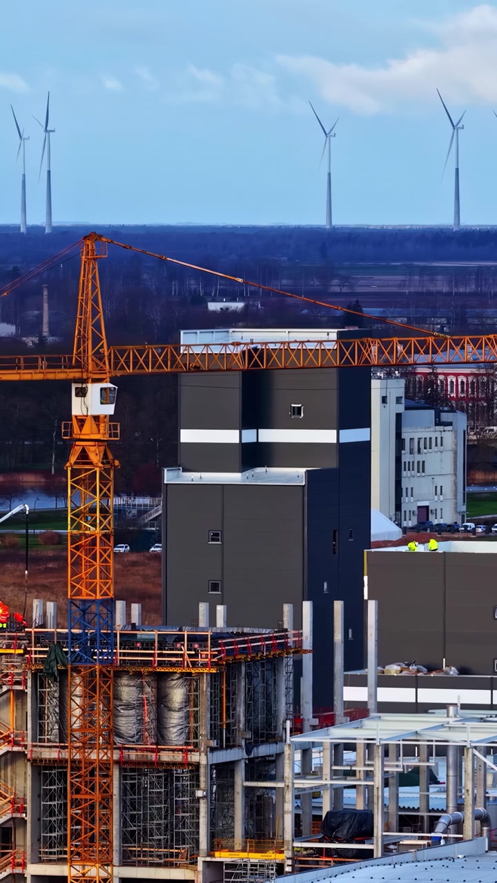 Volo ravvicinato di drone su un grande cantiere industriale in una città rurale innevata nel Nord Europa, con turbine eoliche sullo sfondo, che mostra lo sviluppo energetico moderno in un ambiente freddo