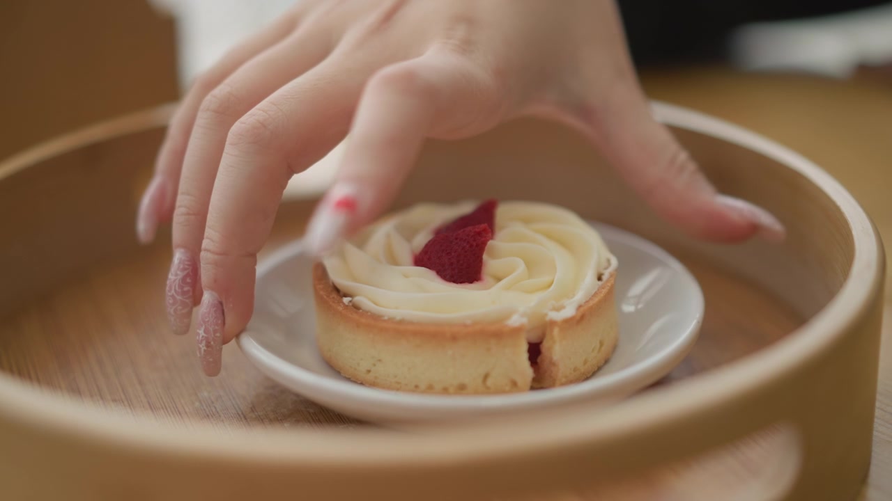 Overhead close view of hand lifting round tart topped with swirled cream and red raspberry garnish from white plate inside circular wooden tray, showing delicate serving action and rich dessert