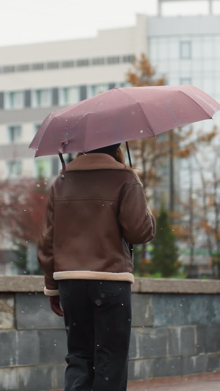 Rear view of young woman walking in light snowfall holding umbrella wearing knit cap brown shearling jacket black trousers one hand pocket autumn leaves urban buildings background
