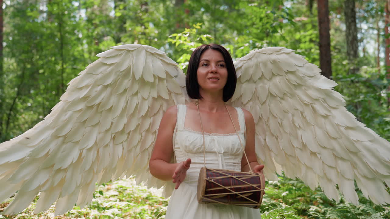 Attrice vestita come una dea con grandi ali d'angelo bianche che cammina con grazia attraverso una lussureggiante foresta verde, sorridendo dolcemente mentre suona la kalimba sotto la luce del sole