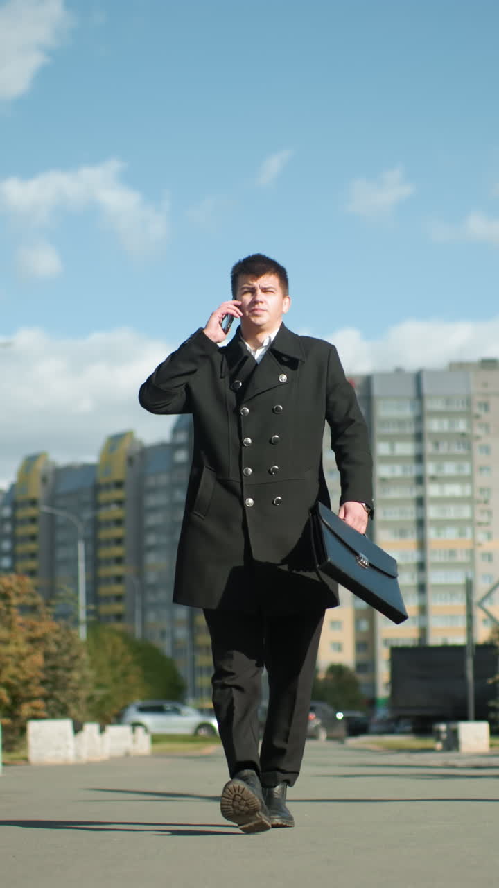 검은 코트를 입은 사업가가 도시 환경에서 외부에서 자신감 있게 고, 들어오는 전화에 응답하기 위해 주머니에서 전화를 꺼내고, 배경에는 초록색, 현대적인 건축물 및 활기찬 도시가 포함되어 있습니다.