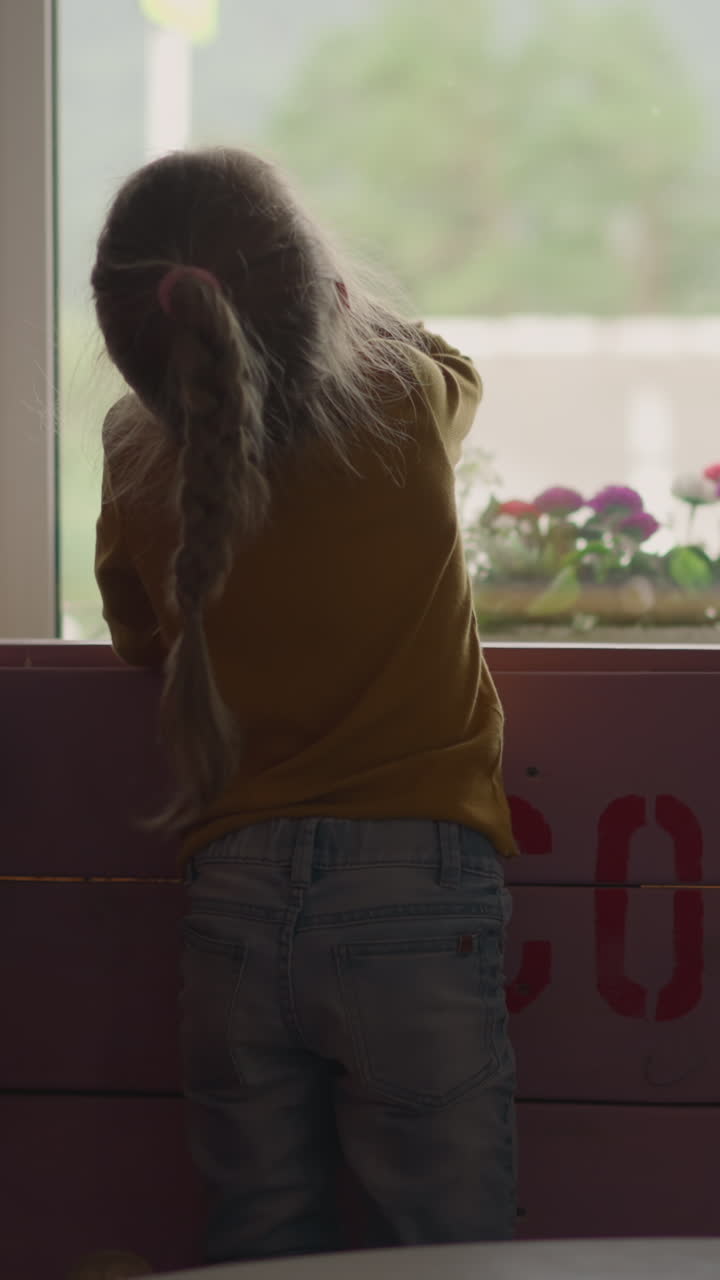 Little girl waves hand and has fun looking out of window with cute younger brother near wooden shield with word Covid-19 in cafe backside view