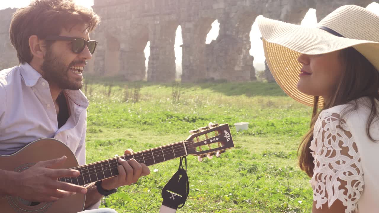 Happy young lovely couple sitting lying on a blanket in the grass in front of ancient roman aqueduct ruins in parco degli acquedotti park in rome romantic play guitar sing beautiful girl with hat bucolic slow motion