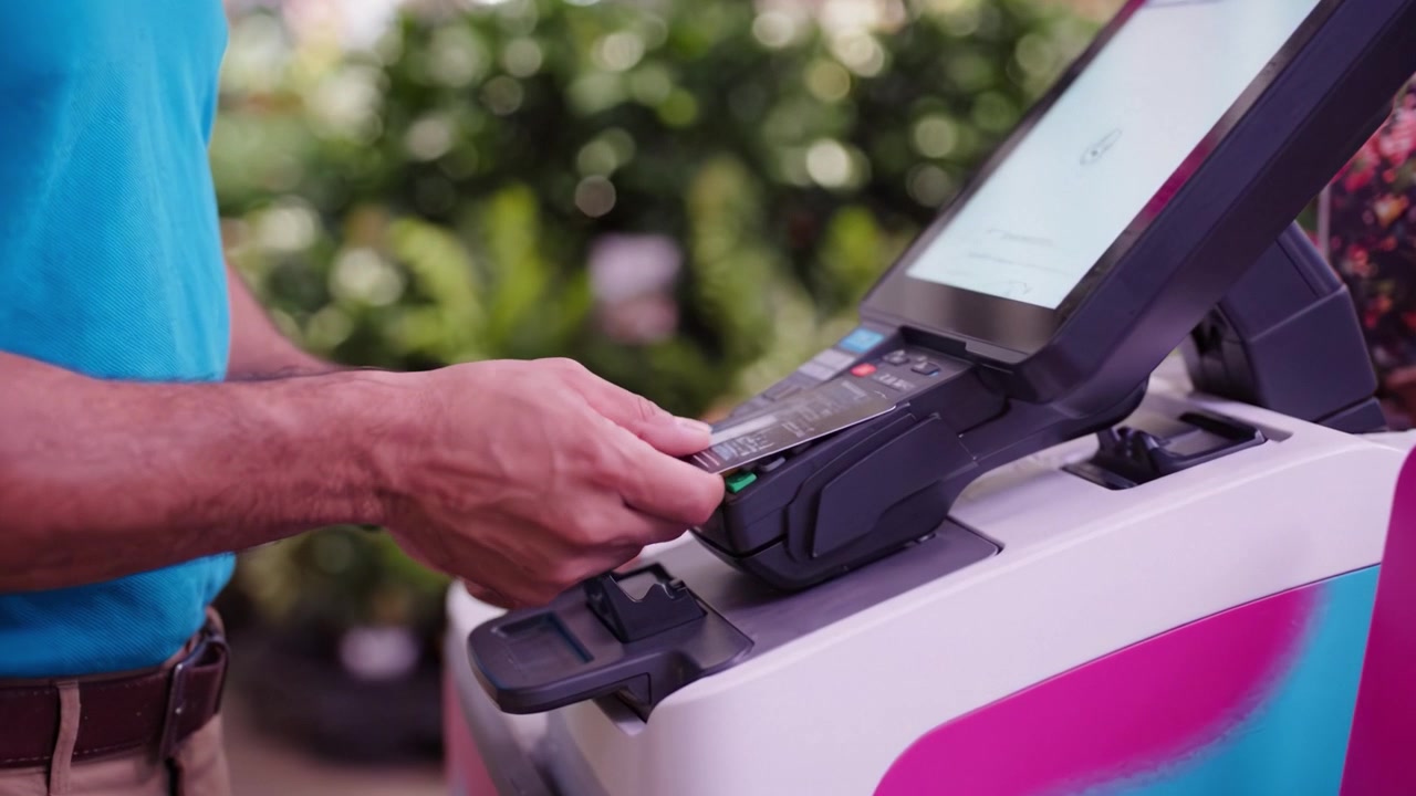 Cliente Usando Auto-pago en un Centro de Jardinería con Exhibiciones Coloridas y Plantas, Procesando el Pago con Tarjeta de Crédito y Dispositivo Móvil en la Caja.