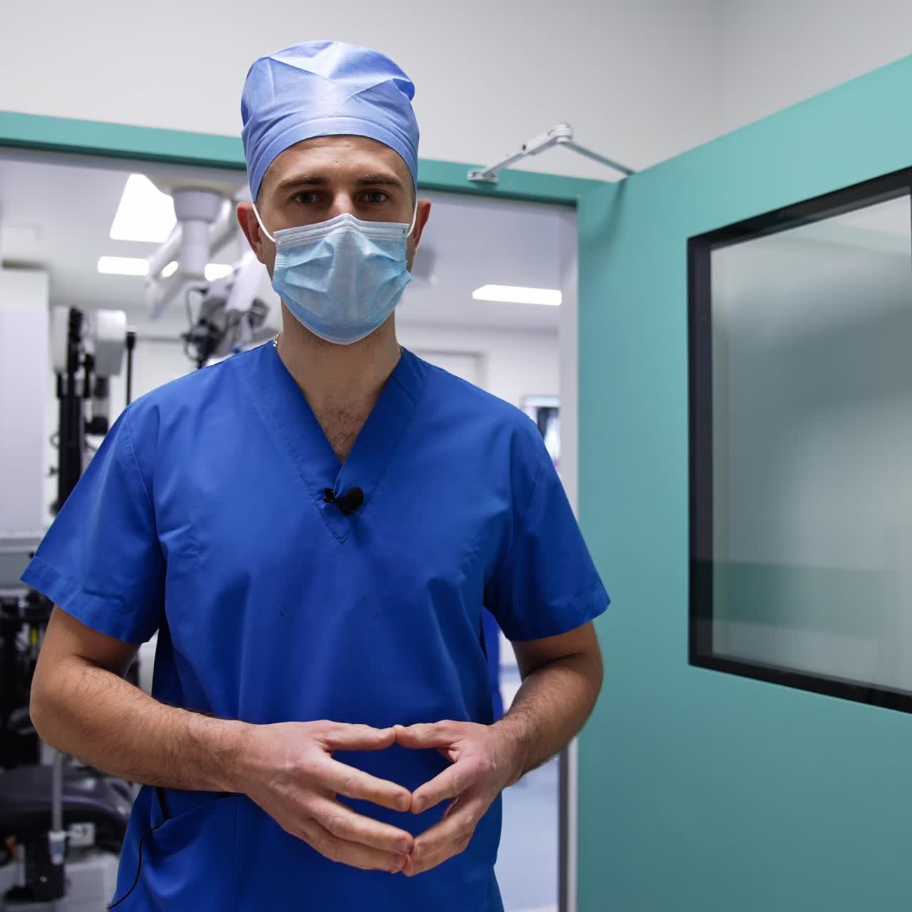 Adult male surgeon in mask and special clothes talks to the camera. Medic stands at the doors of surgery room and then closing it