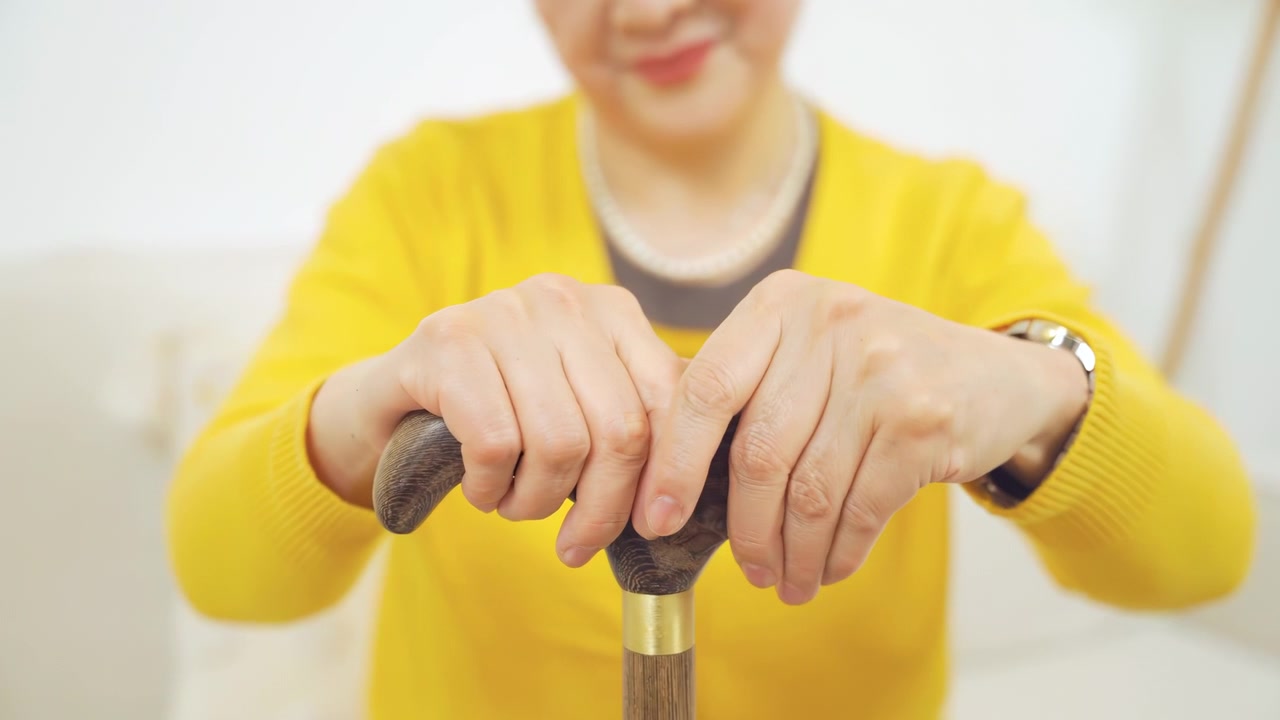 Anziana donna sorridente con bastone da passeggio, la testa appoggiata sulla mano, che indossa un abbigliamento casual, seduta su un comodo divano; la nonna tiene il pomello del bastone, soffre di problemi muscolari e malattie ossee.