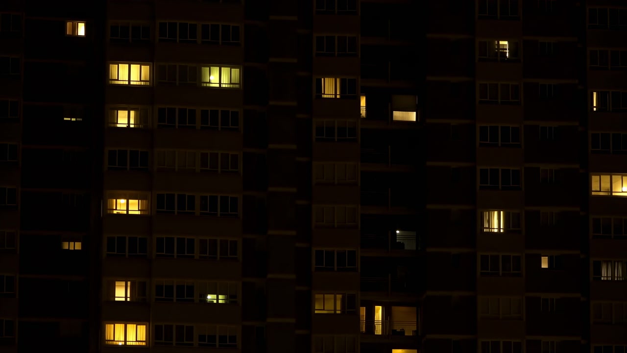 Esterno notturno della facciata di un edificio residenziale dove riflessi circolari sfocati e tenui bagliori delle finestre si trasformano gradualmente in rettangoli illuminati più nitidi, rivelando ambra calda e verde pallido.