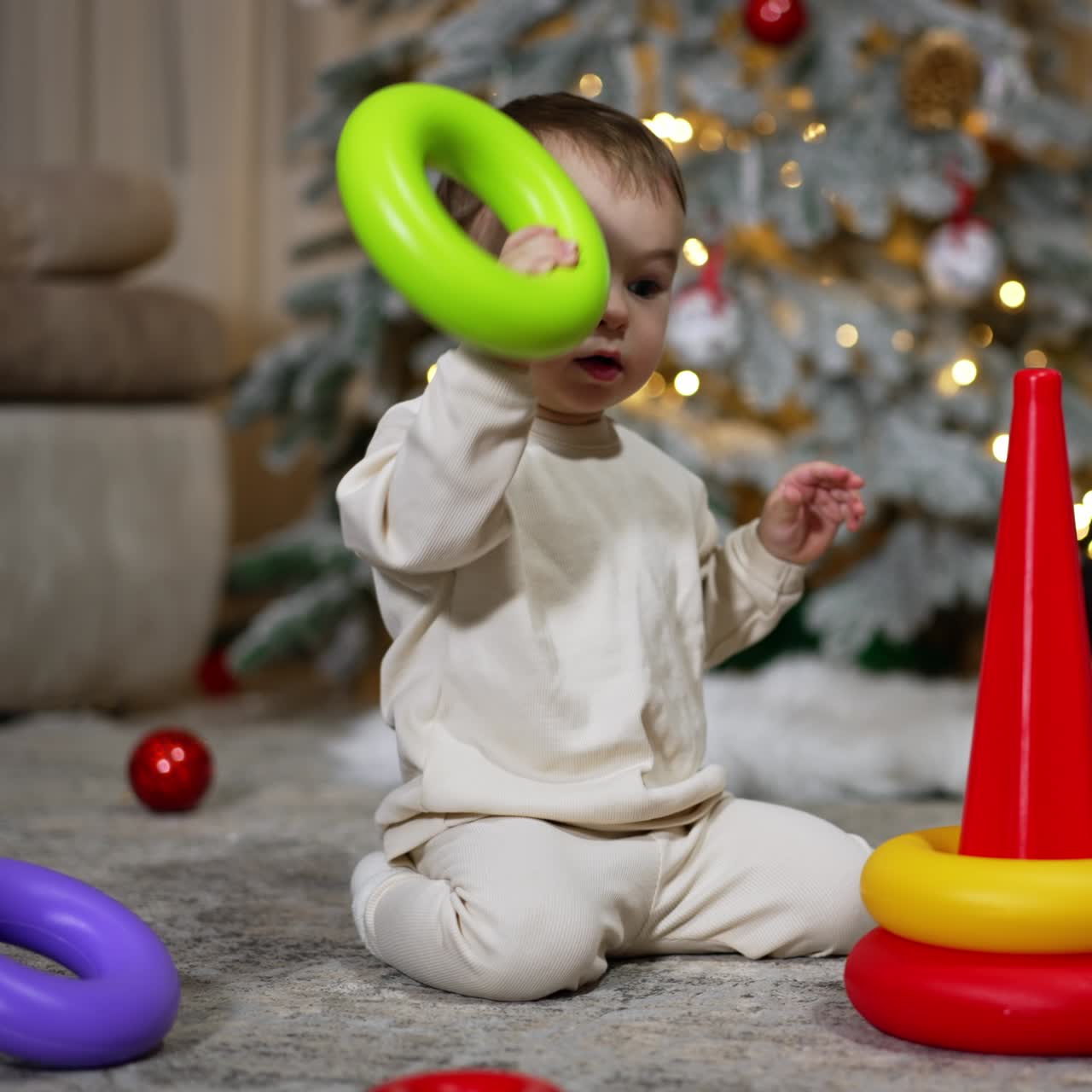 Cute little Caucasian toddler playing with a pyramid sitting on the floor. Christmas tree with a black cat sitting beside at backdrop in blur