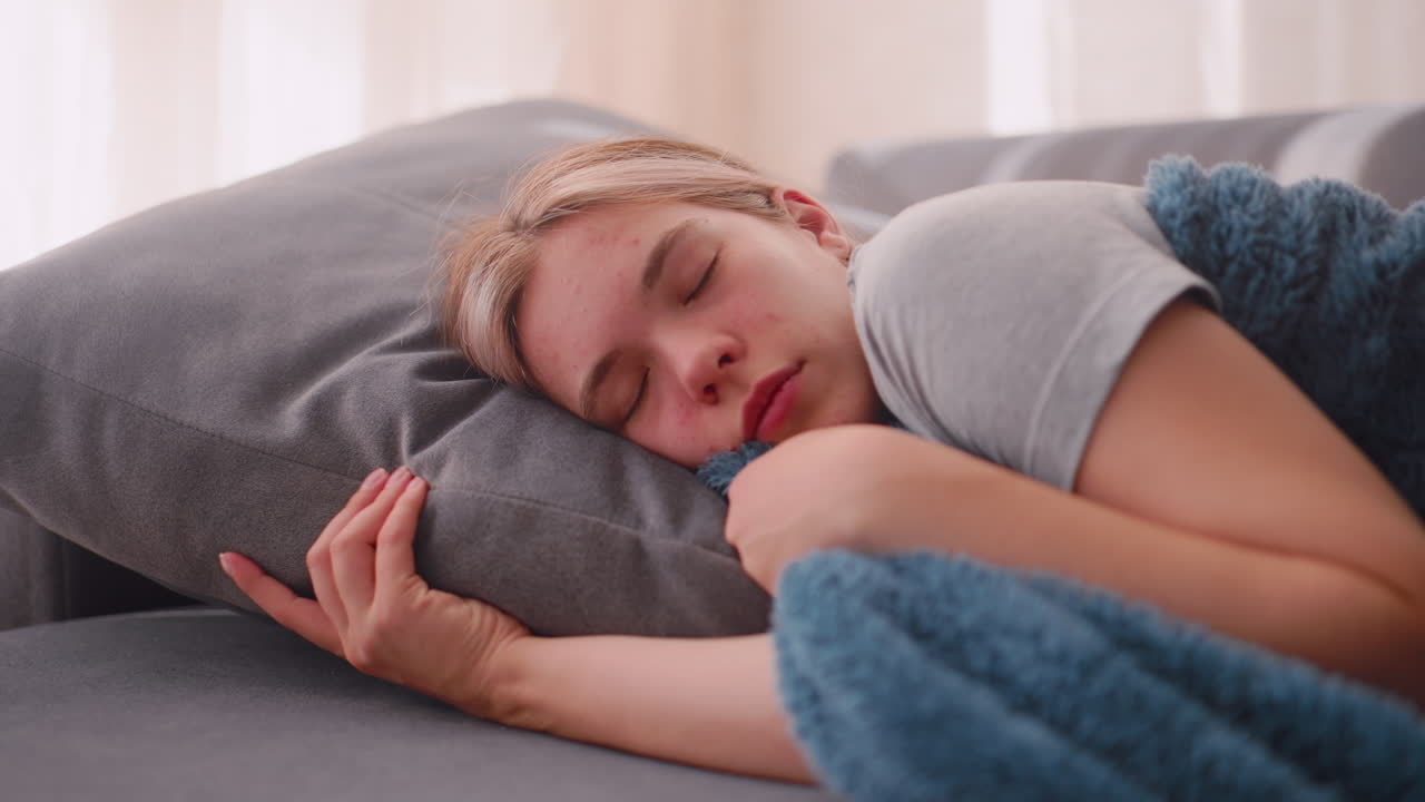 Dreamer peacefully holding blanket warmly while sleeping on couch with relaxed face and closed eyes enjoying comfort in soft daylight streaming through curtains creating serene atmosphere of rest