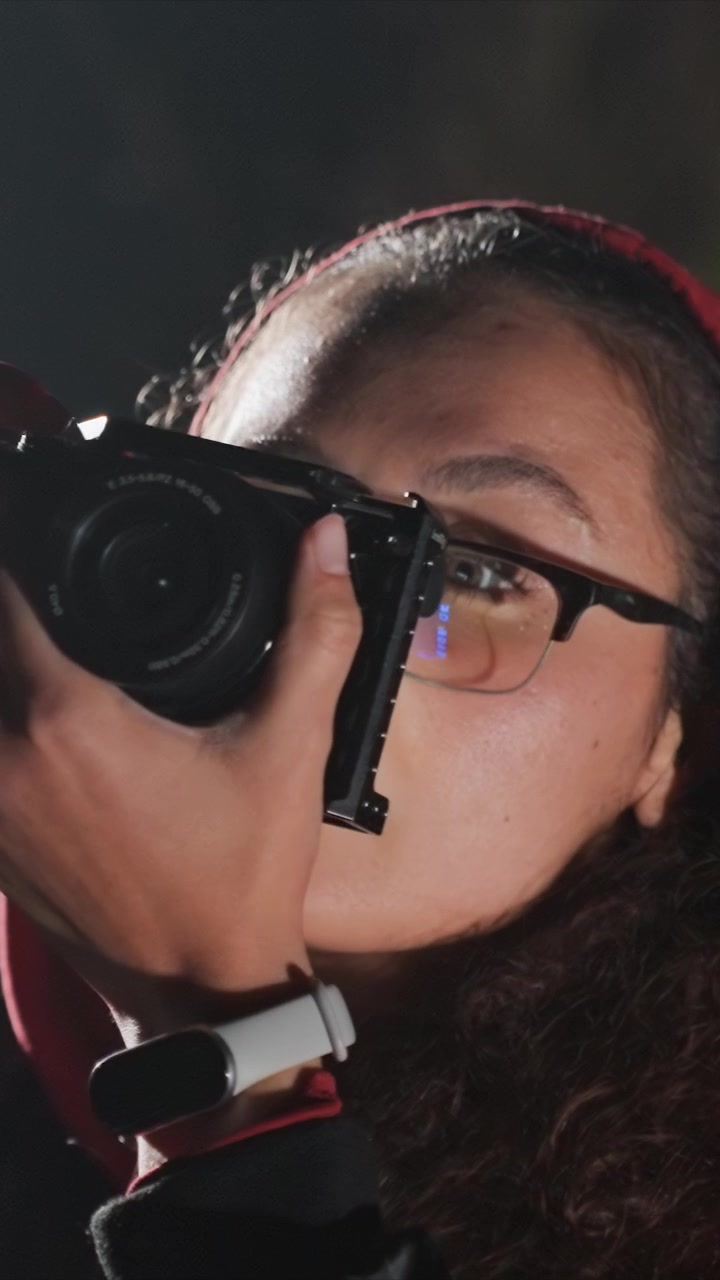 Curly haired woman in red hoodie adjusting cameras. Reflections appear in her glasses as she manipulates camera. Close up composition emphasizes clothing and skin details