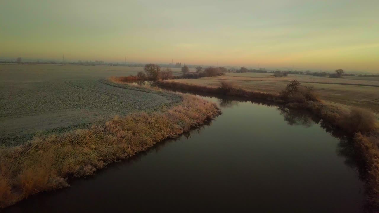 Drone flyver lavt over en bugtende flod og frostklare marker med solopgang bag dronen